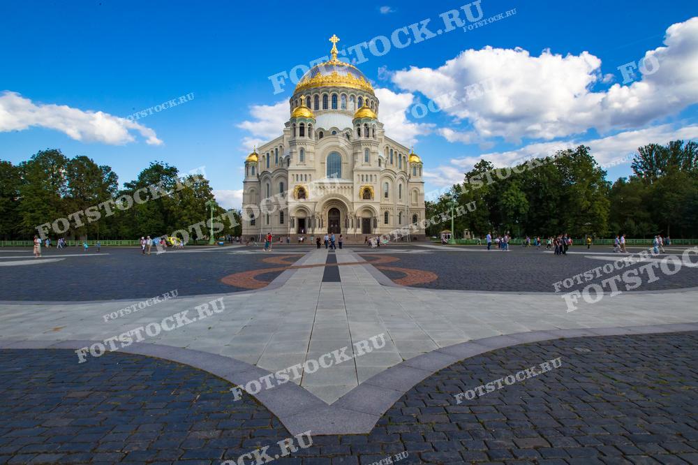 На фотографии изображен Морской собор в Кронштадте, расположенный на площади Якорной. Главный фасад собора украшен золотыми куполами, сверкающими на солнце. Вокруг собора находятся зеленые деревья и просторная площадь, где гуляют люди. Летний день с ясным небом подчеркивает величие архитектуры.